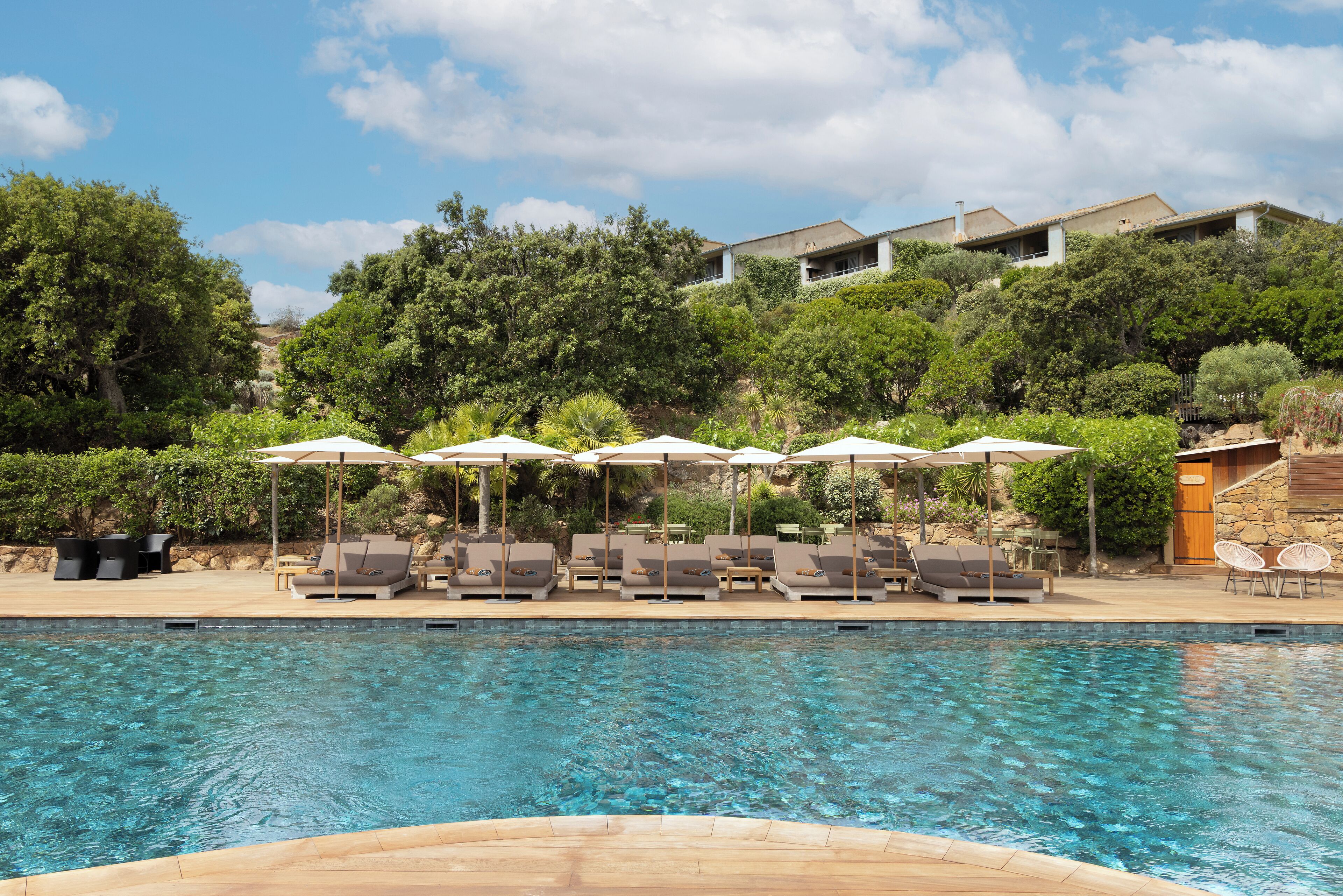 Une piscine entourée de verdure, avec des chaises longues et des parasols, et un bâtiment situé sur une colline à proximité.