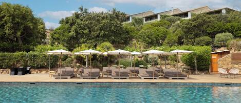 A swimming pool with lounge chairs and umbrellas, surrounded by greenery and a hillside building.