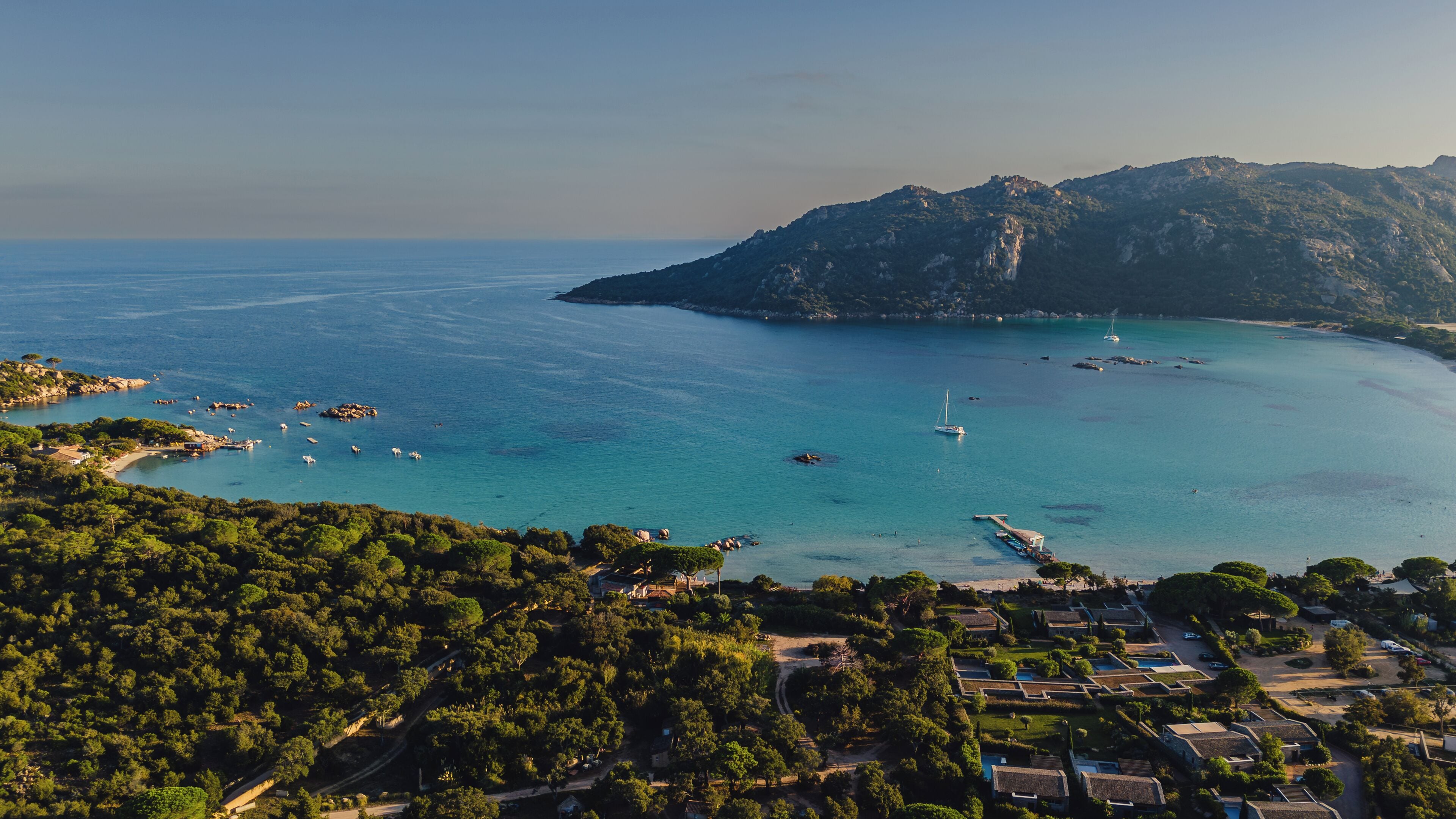 An aerial view of a coastal area with clear blue waters, boats, and a mountainous backdrop.