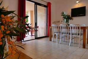 Dining area with a dining table and four chairs, adjacent to a room with a flat-screen TV and a potted plant.