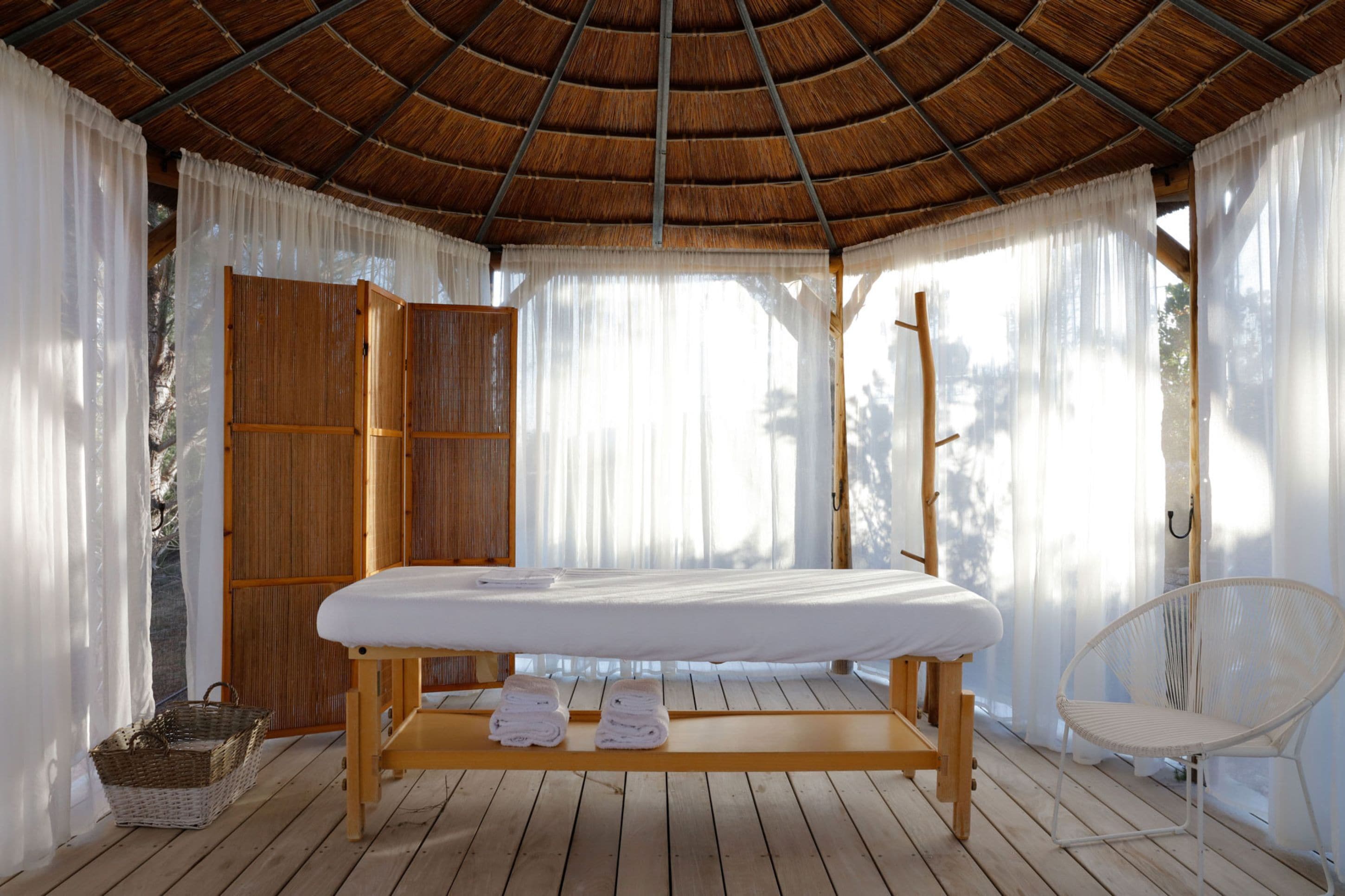 A spa room with a wooden massage table, white towels, a wicker basket, and a white chair.