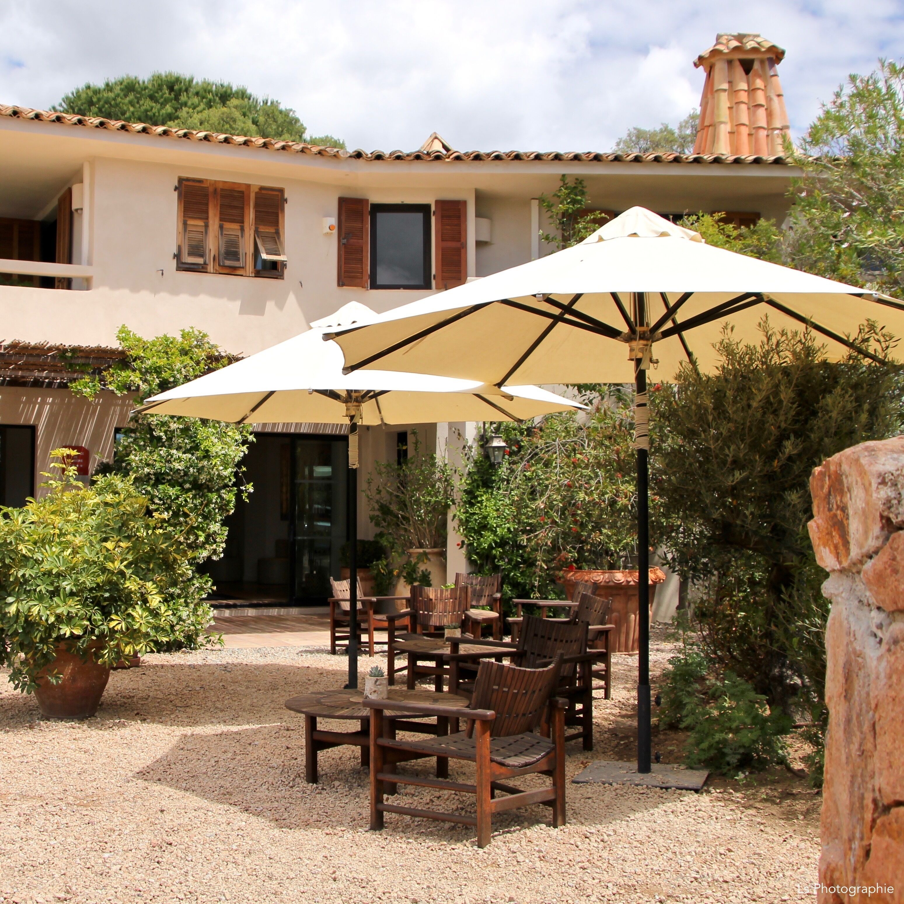 A patio area with outdoor furniture and umbrellas, surrounded by potted plants and a building with a tiled roof.