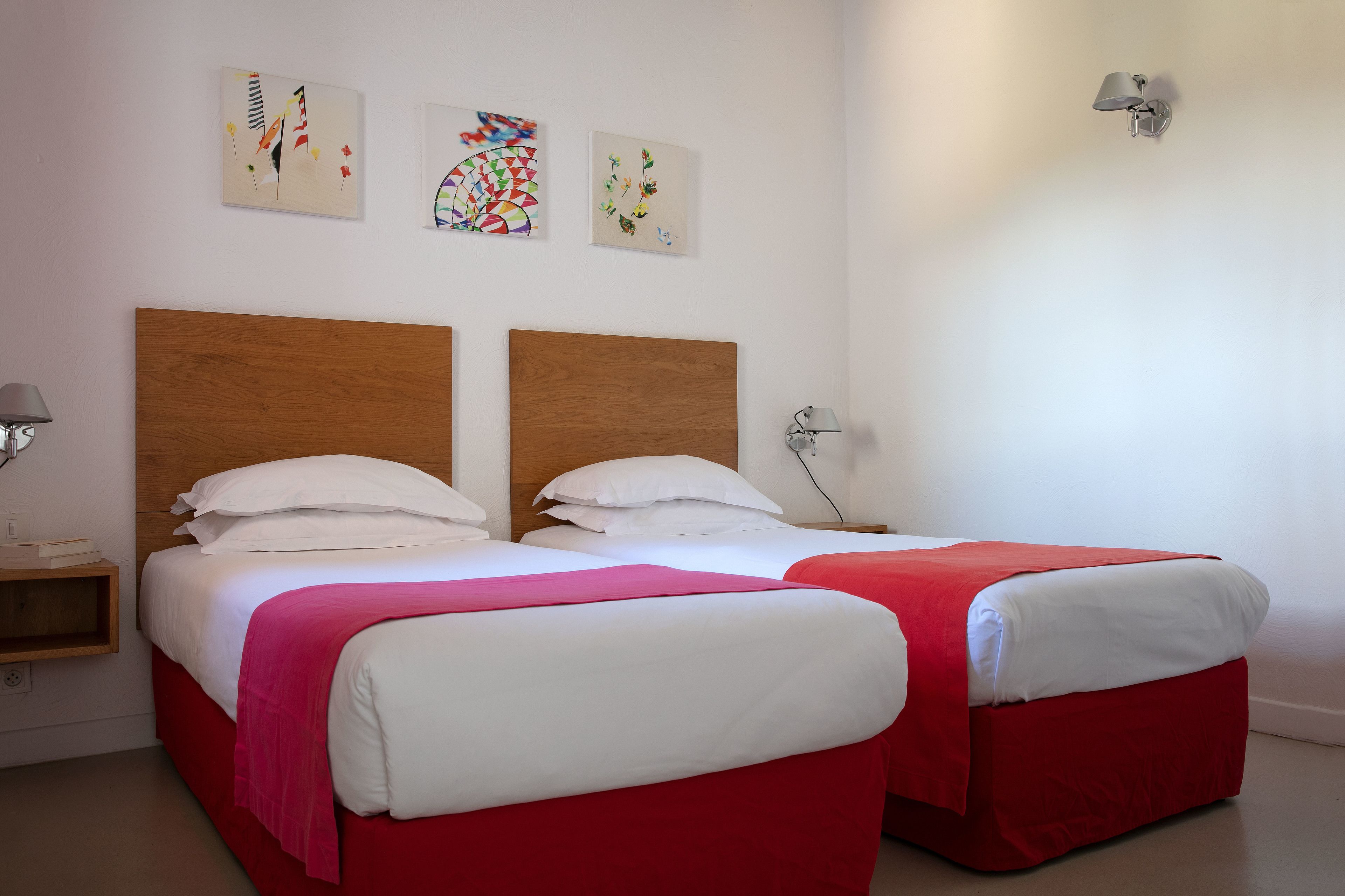 A hotel room with two beds, each with a wooden headboard, white bedding, and red bed skirts. There are three artworks on the wall, a nightstand with a lamp, and a wall-mounted light fixture.