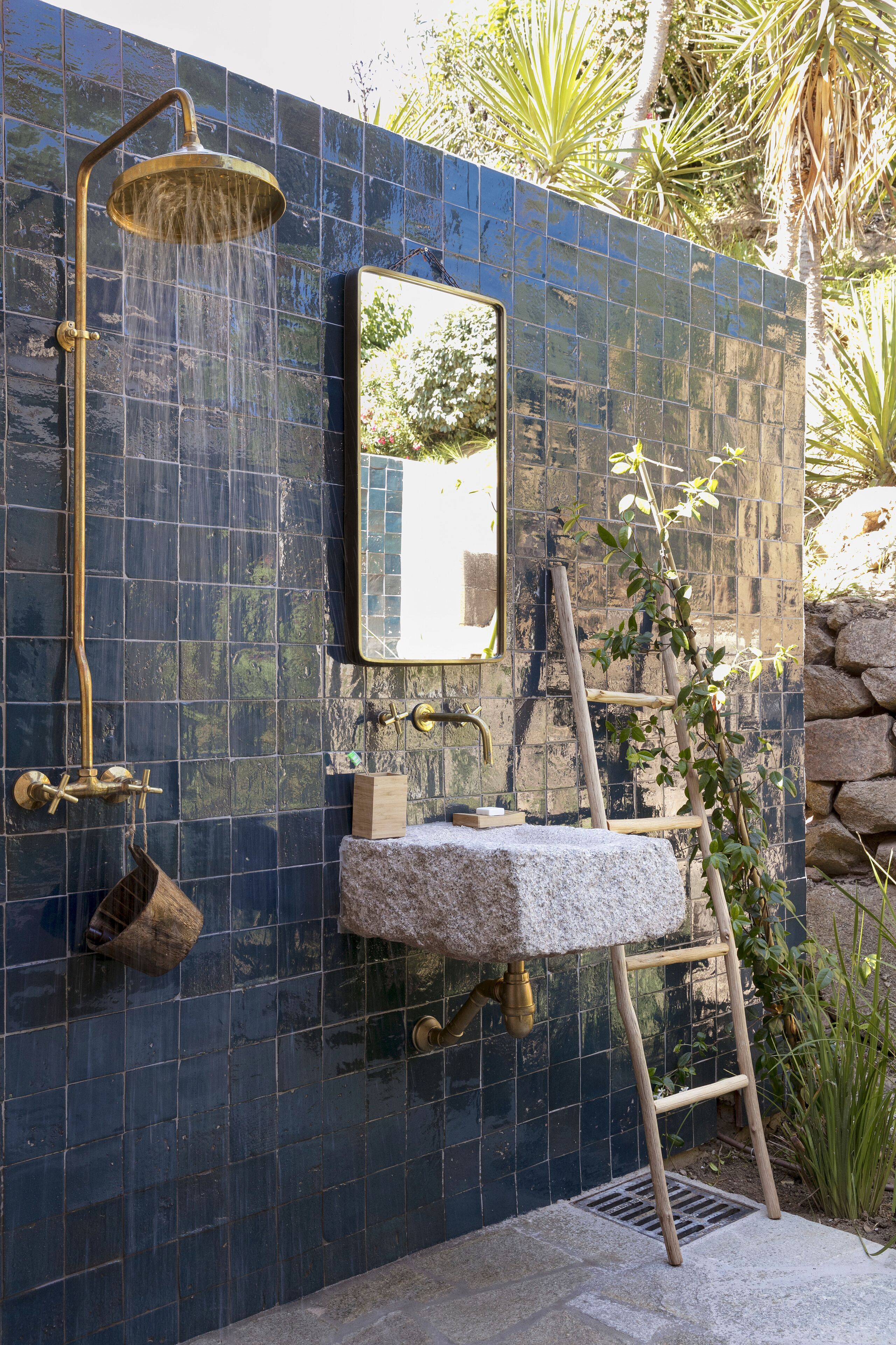 A bathroom with a blue tiled wall, a mounted mirror, a stone sink, and a shower with a brass fixture.