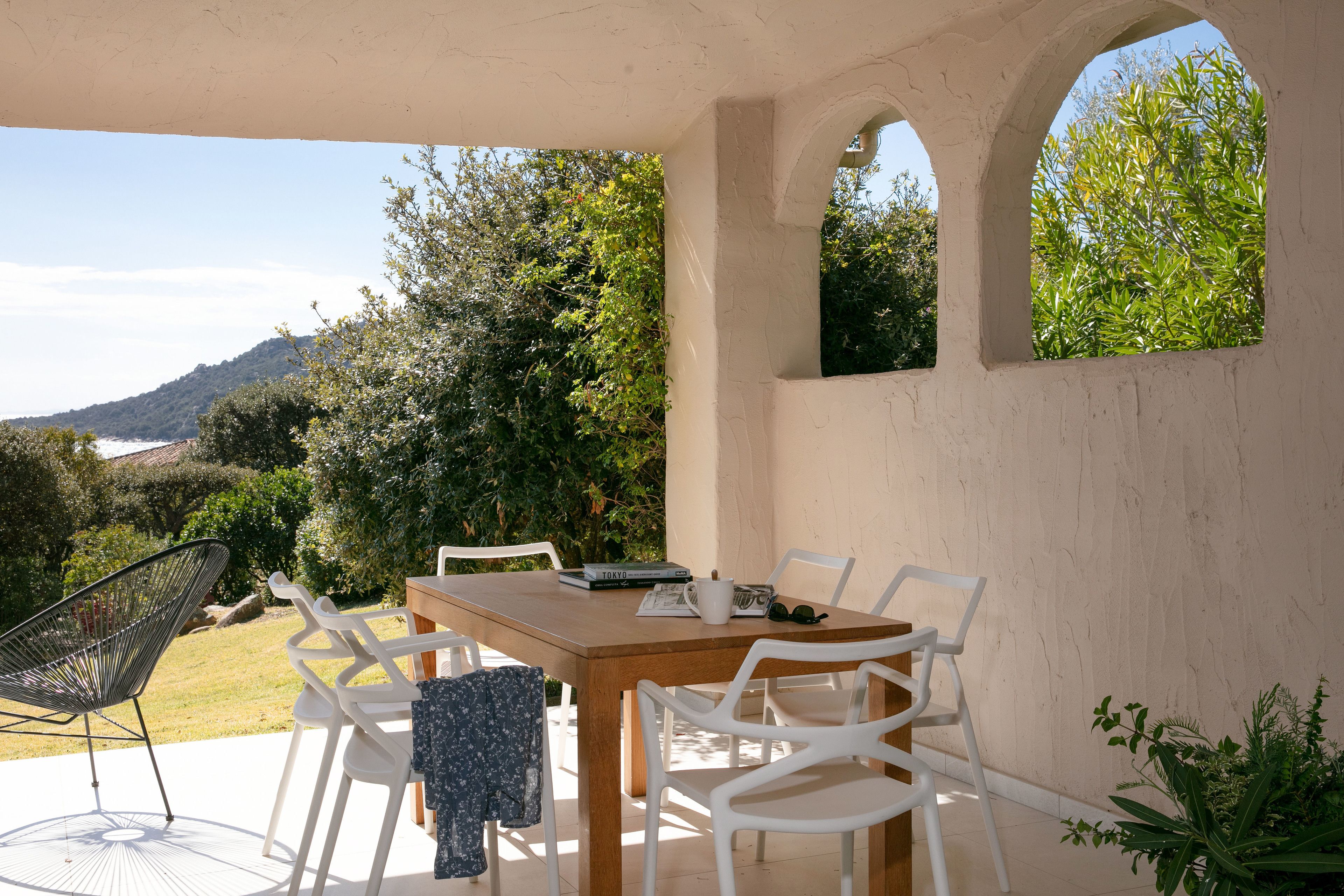 A covered outdoor dining area with a wooden table and white chairs, overlooking a lush garden and distant hills.