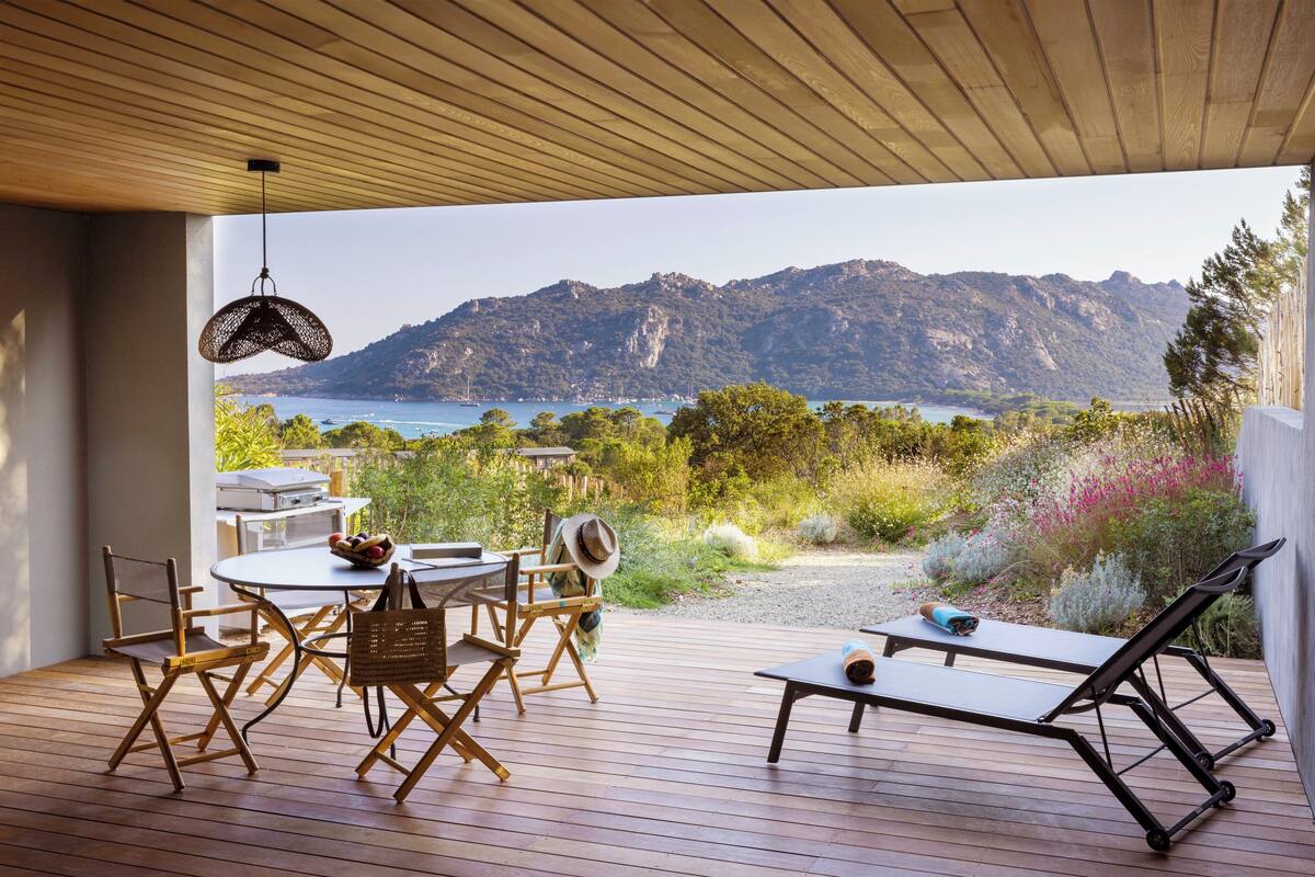Une terrasse extérieure dotée d’un plafond en bois, d’une table à manger avec des chaises, d’un fauteuil de relaxation et offrant une vue sur un paysage montagneux et la mer.