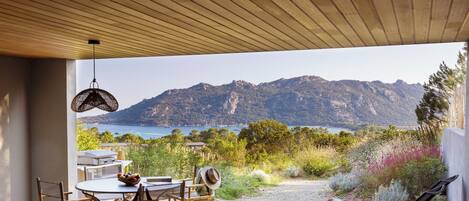 An outdoor deck with a wooden ceiling, a dining table with chairs, a lounge chair, and a view of a mountainous landscape and sea.