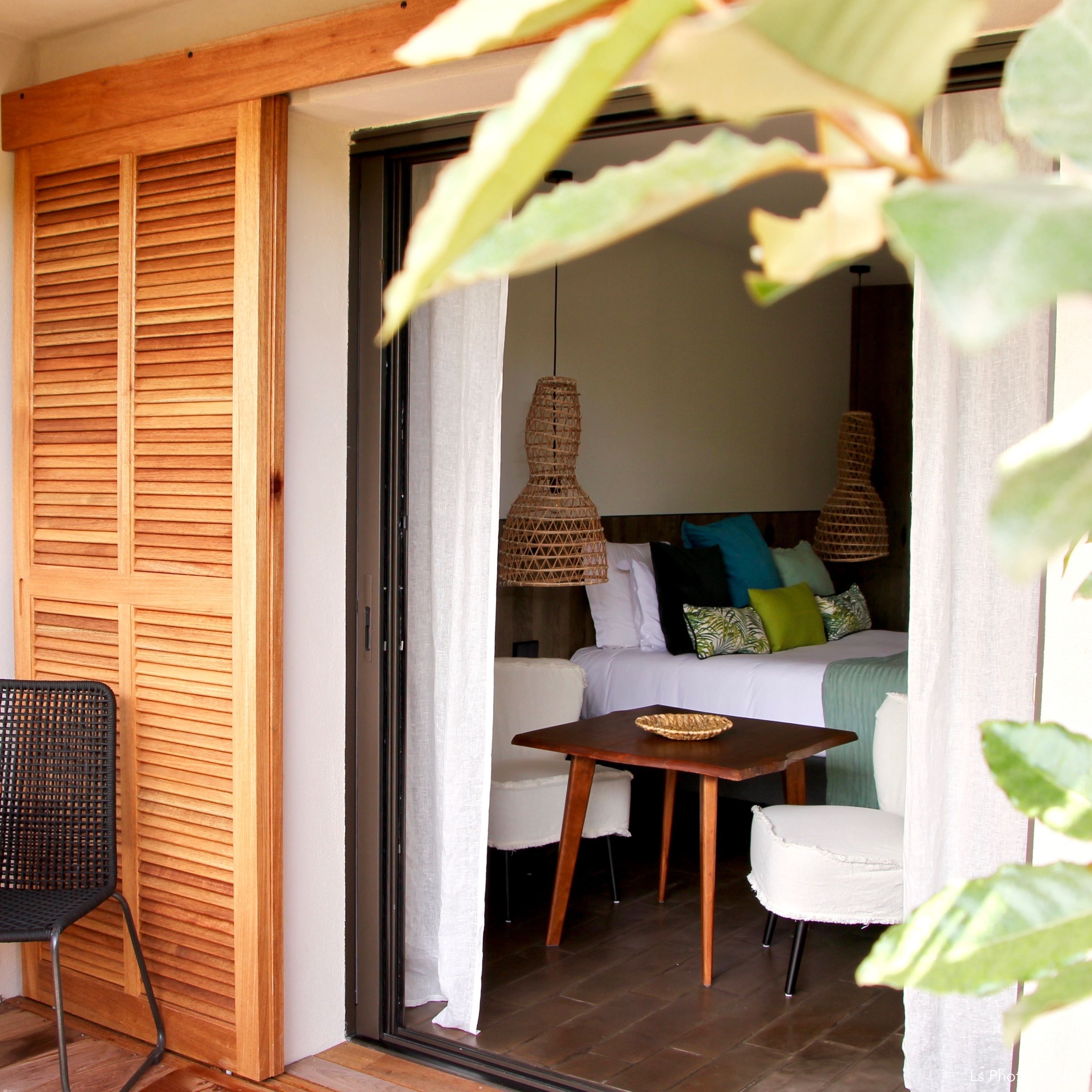 A room with a wooden slatted door, a wicker chair, a small table with a woven bowl, a bed with colorful pillows, and a hanging light fixture.