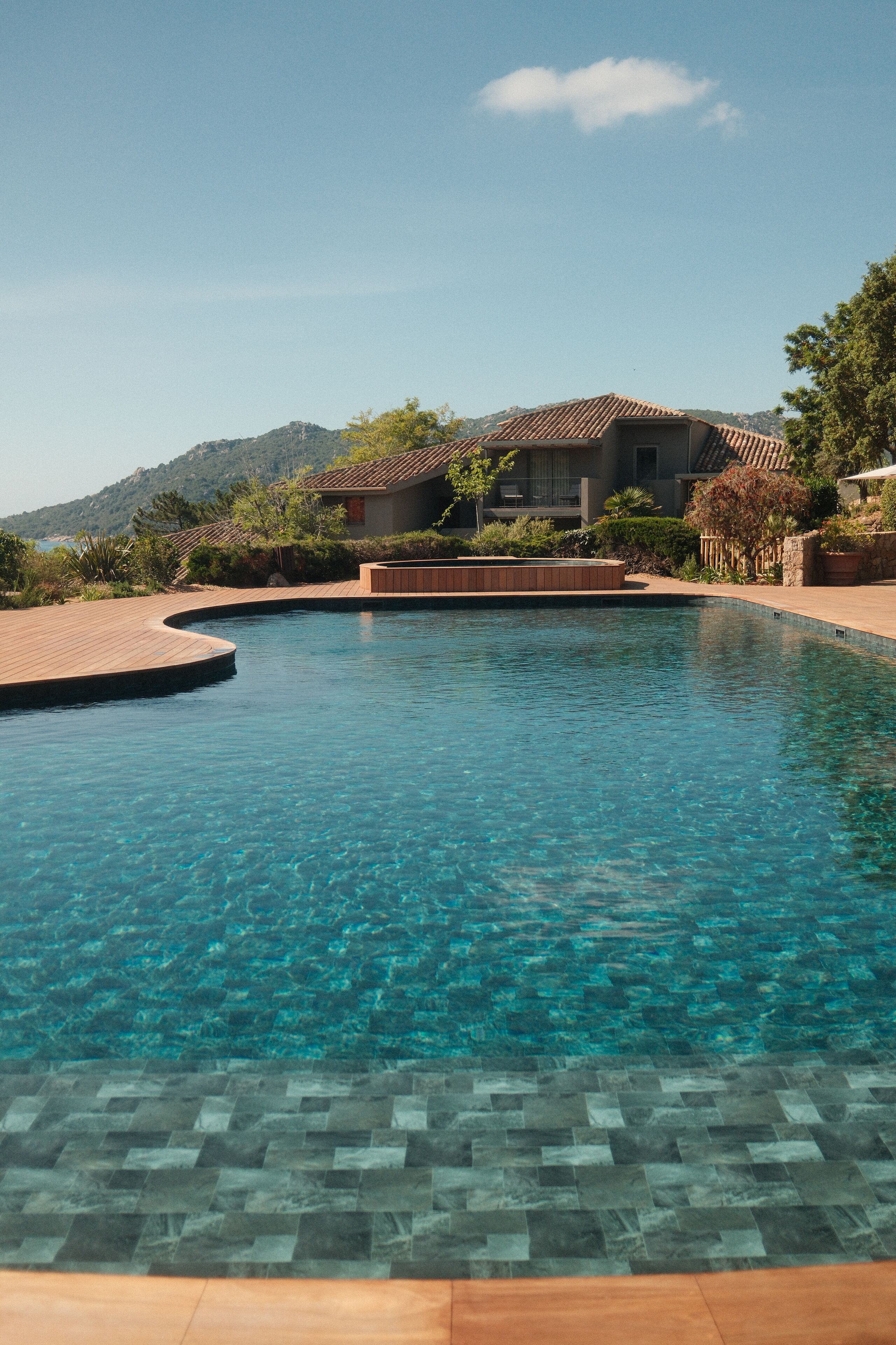 A clear swimming pool with a tiled deck, surrounded by a landscaped garden and a building with a tiled roof.