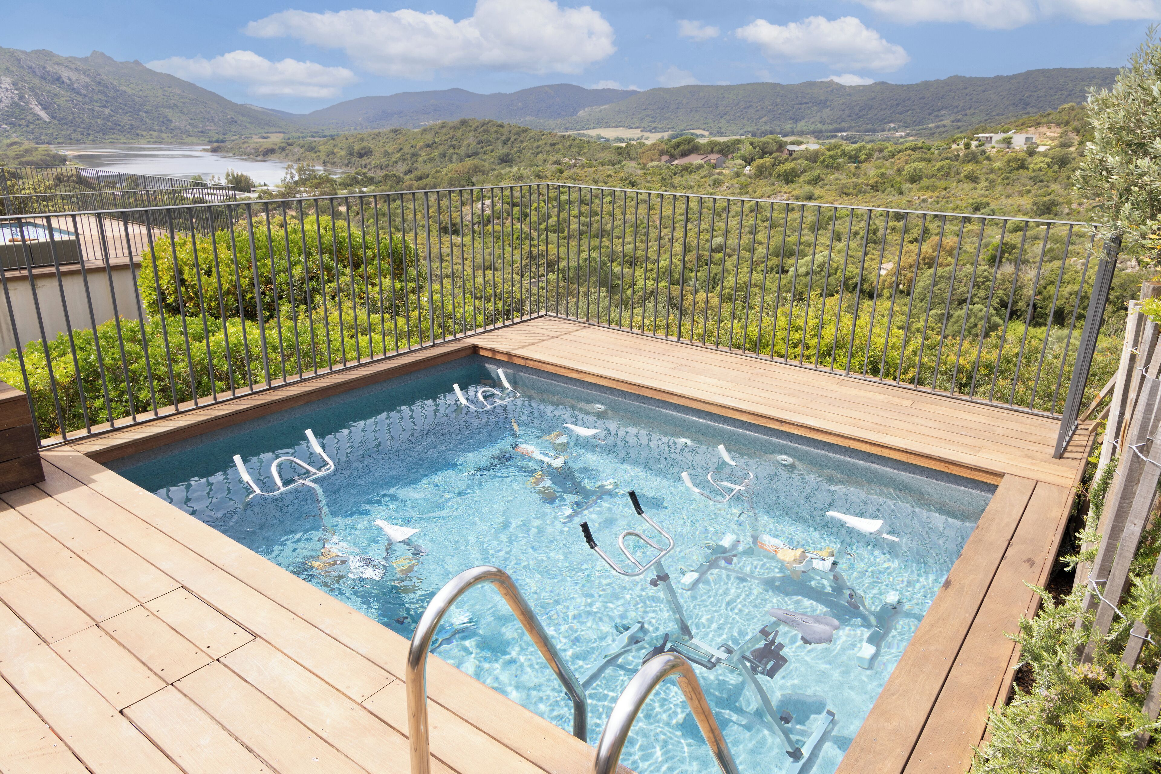 A rectangular pool with a wooden deck and metal railings, surrounded by lush greenery and a mountainous backdrop.