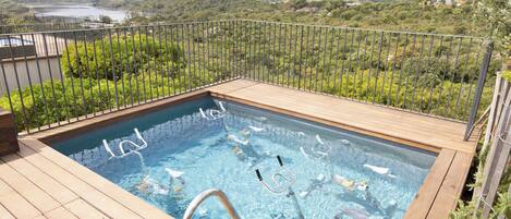 A rectangular pool with a wooden deck and metal railings, surrounded by lush greenery and a mountainous backdrop.