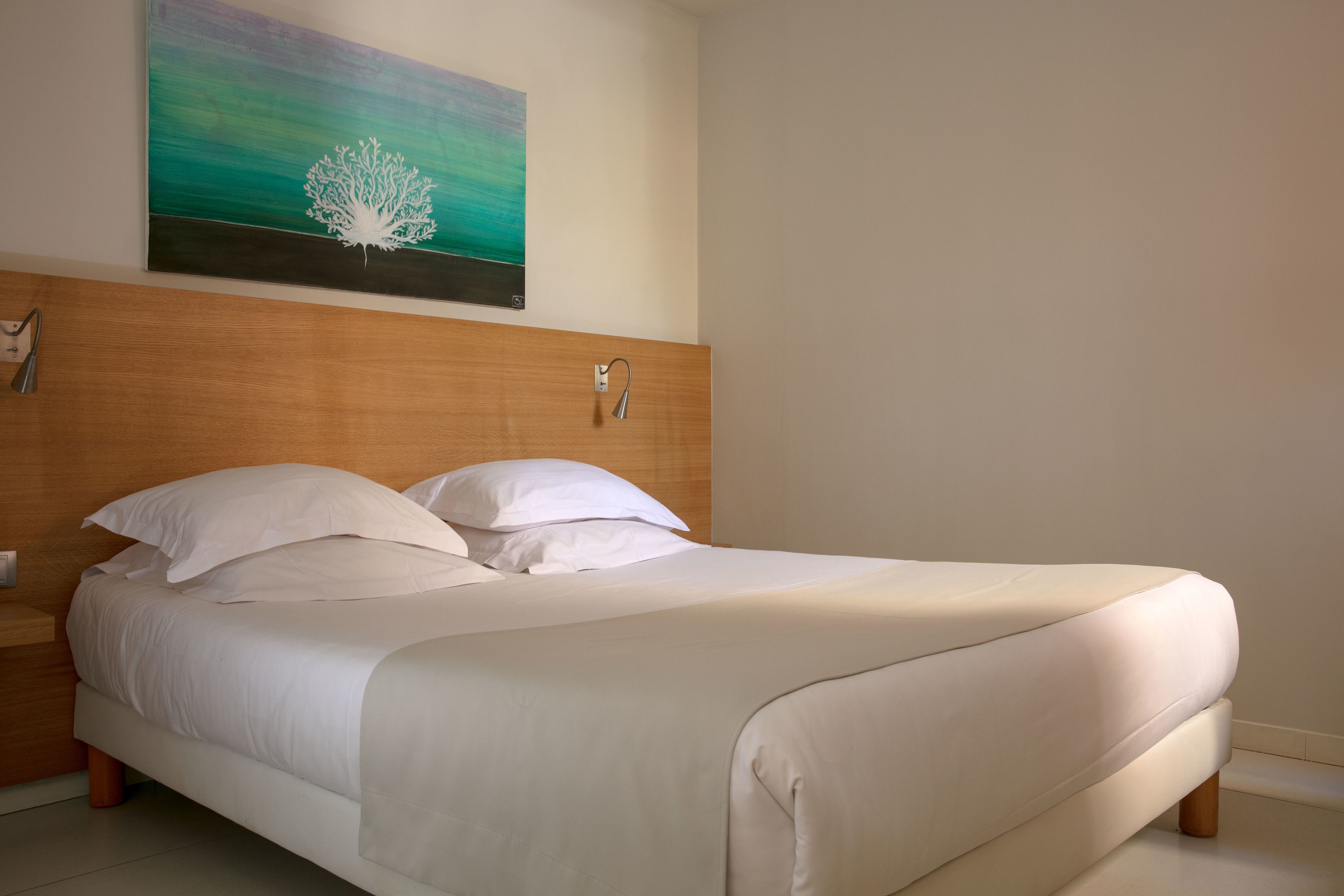 Un lit bien fait, avec du linge de lit blanc et des oreillers, une tête de lit en bois et un tableau représentant un arbre accroché au mur.