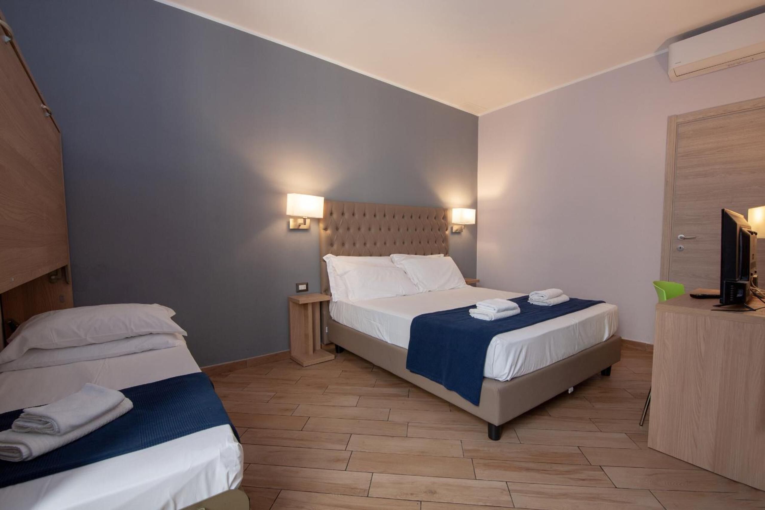 Ein Hotelzimmer mit zwei Betten, Holzboden, einem Kopfteil mit Wandleuchten, einem Schreibtisch mit Stuhl und einer Tür.
