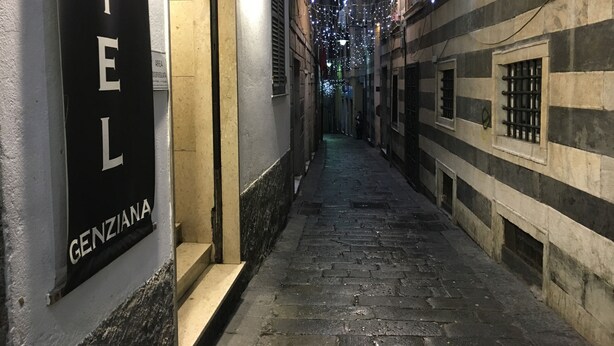 Une ruelle étroite, avec une enseigne d’hôtel indiquant « HÔTEL GENZIANA » sur le côté gauche. La ruelle est bordée de bâtiments des deux côtés, et des guirlandes lumineuses sont suspendues au-dessus.