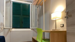 Une petite pièce avec un bureau en bois, une chaise verte, une fenêtre avec des stores, un ventilateur sur pied et une lampe fixée au mur.