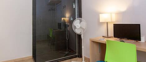 Habitación de hotel con cabina de ducha de vidrio, un ventilador de pie, un escritorio con una computadora, una silla verde y un basurero azul.