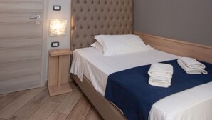 Un lit bien fait, avec du linge de lit blanc et une couverture bleue, une table de chevet en bois avec une lampe, et une porte dotée d’un trou de serrure.