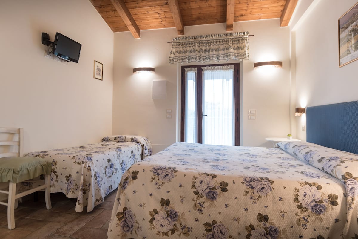 A room with two beds, a television mounted on the wall, a small table, and a window with curtains.