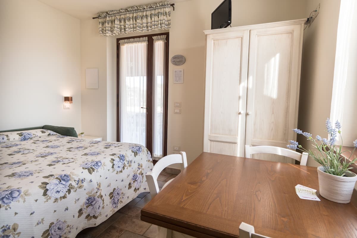 A bedroom with a floral bedspread, a wooden dining table with chairs, a potted lavender plant, and a wardrobe.