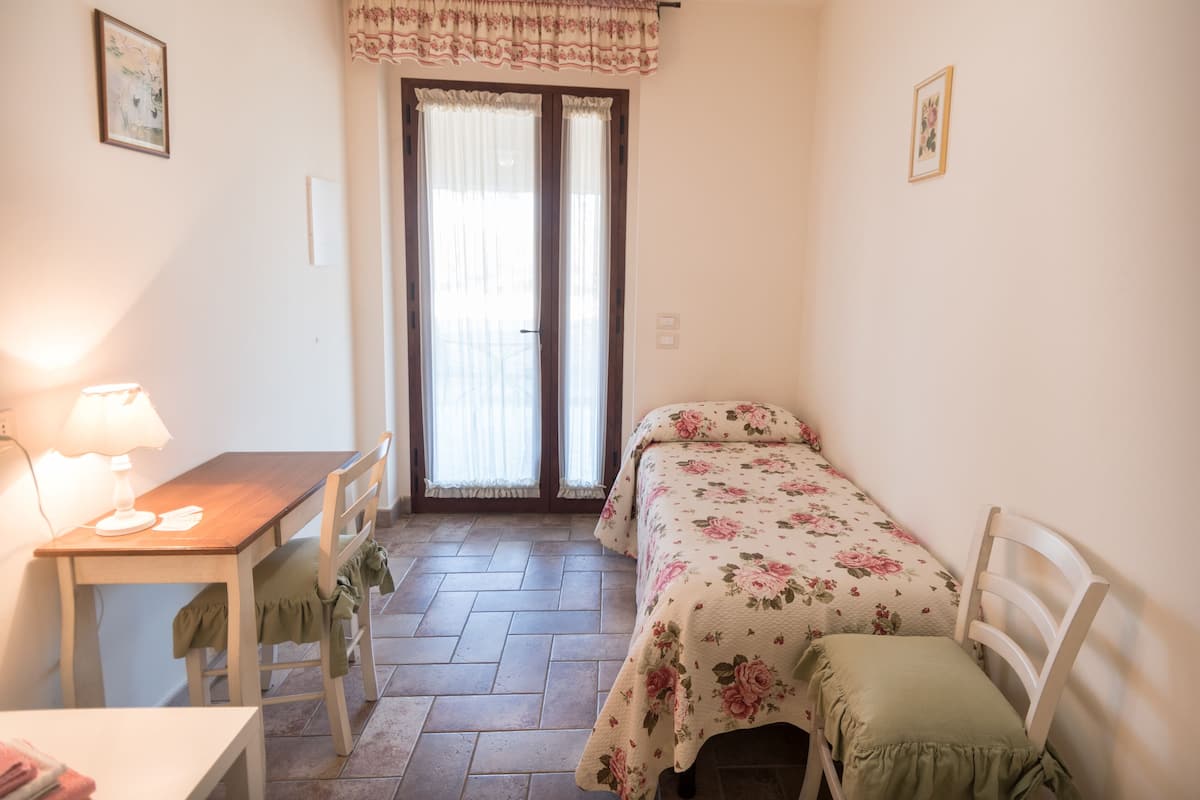 A small, cozy room with a wooden desk, a bed with floral bedding, a chair, and a window with curtains.