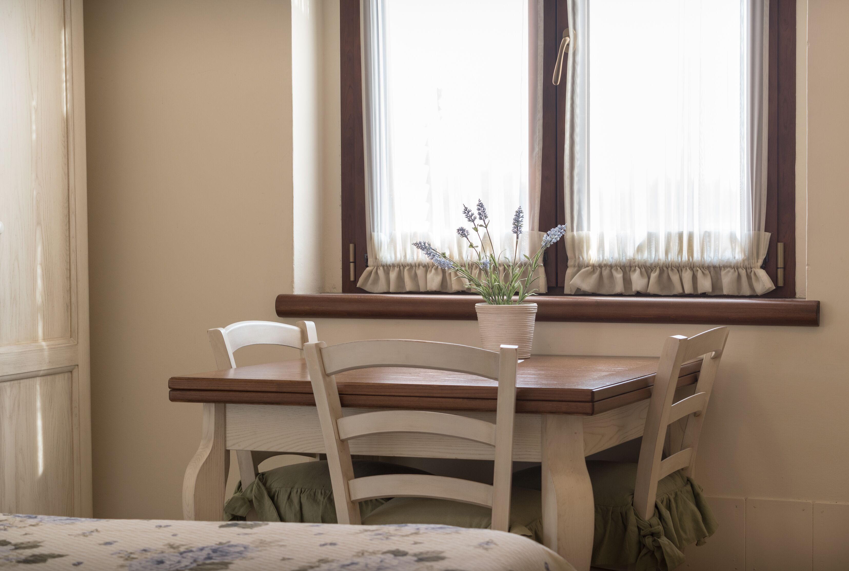 Un coin repas avec une table en bois et deux chaises blanches, un pot de lavande posé sur le rebord de la fenêtre et des rideaux légers aux fenêtres.