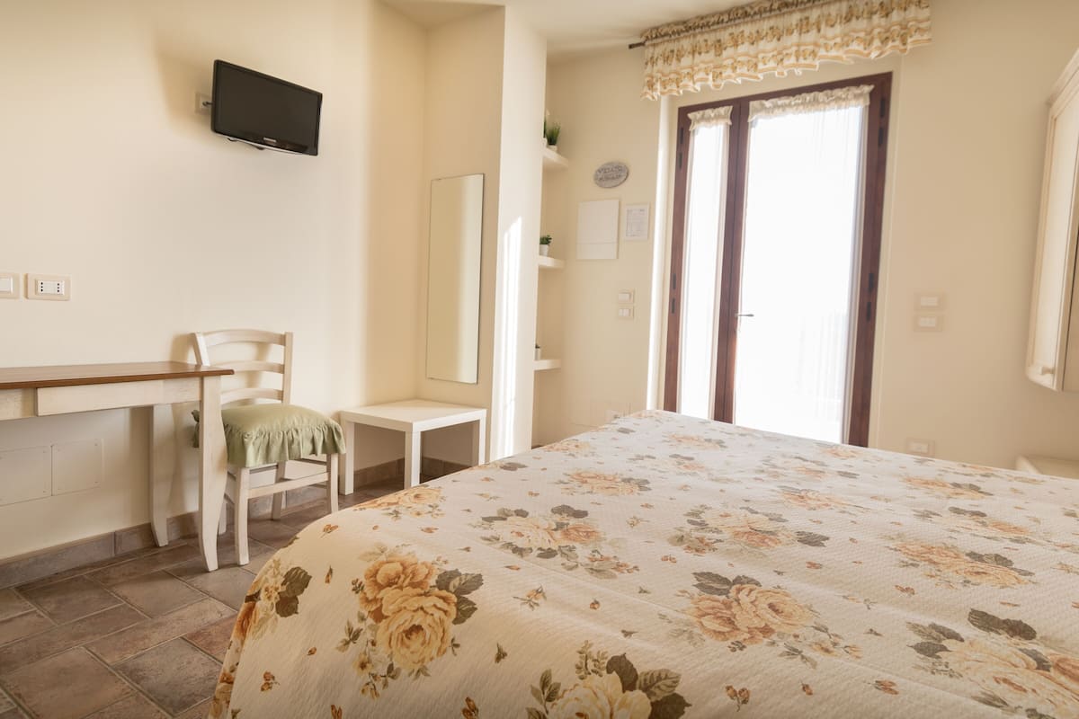A bedroom with a bed, a desk, a chair, a television, a window with curtains, and a door.