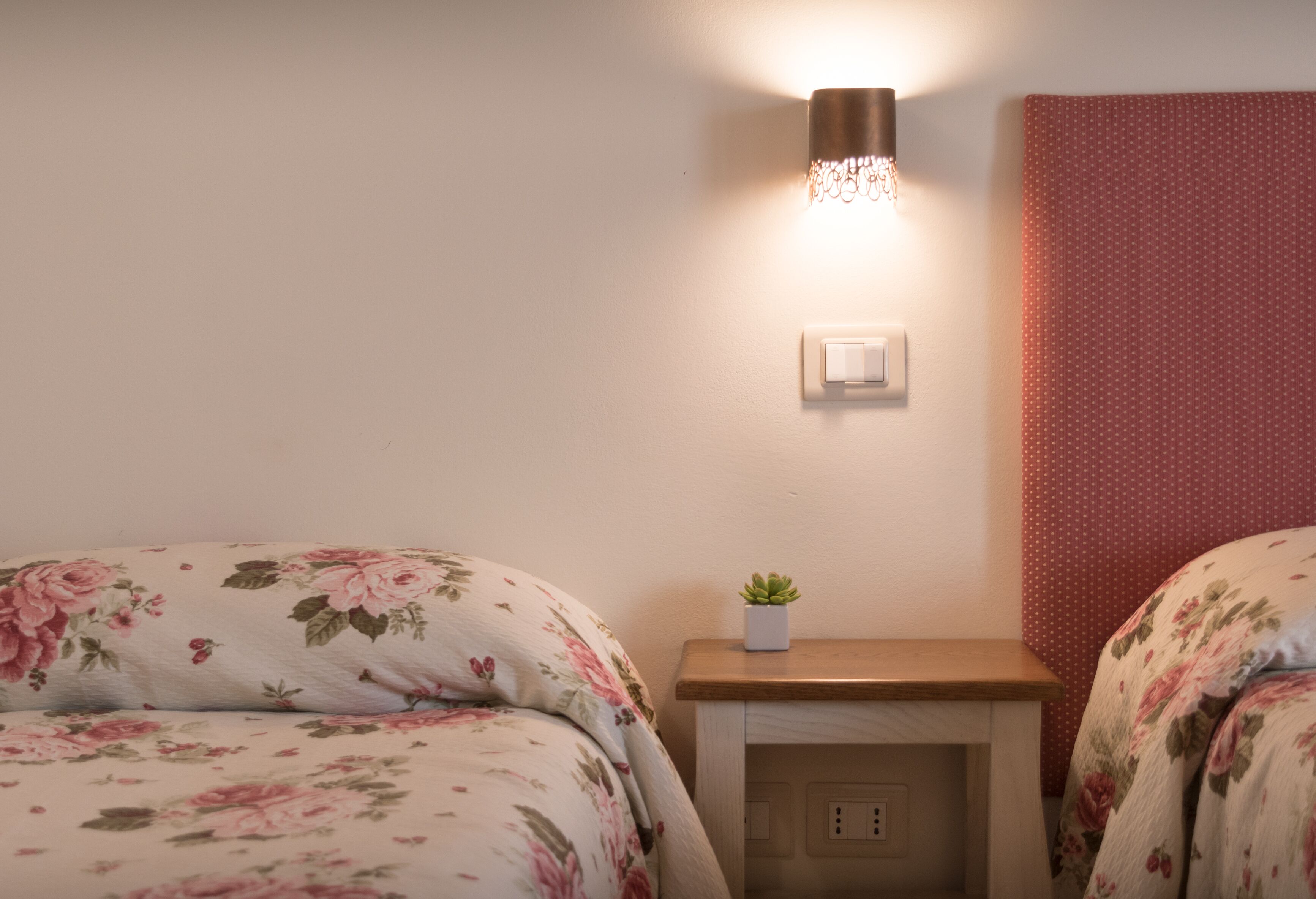 Un dormitorio con ropa de cama floral, una mesita de noche con una planta, una lámpara de pared y un enchufe.