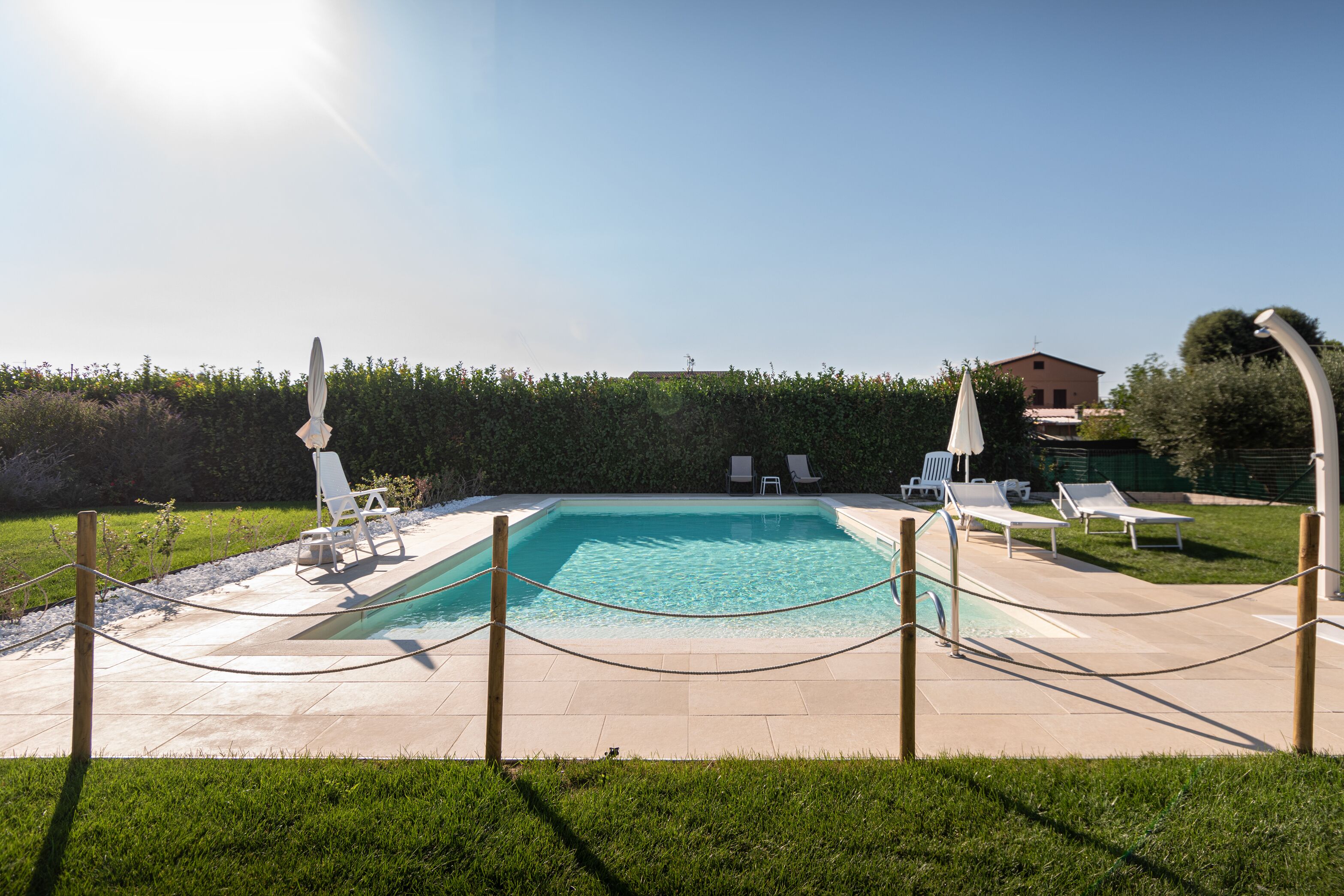 A rectangular swimming pool with a clear blue water, surrounded by a low stone wall and a wooden fence. There are several white lounge chairs and umbrellas around the pool.