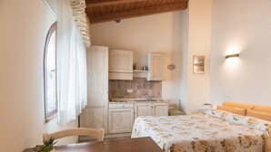A cozy bedroom with a bed, a wooden table with a plant, and a view of a kitchen with cabinets and a window. - Rosa di Assisi (Bastia Umbra)