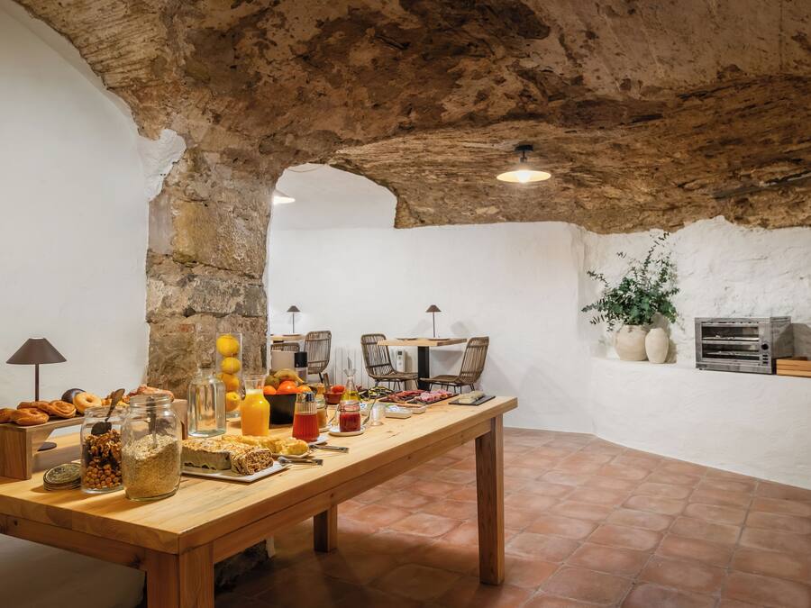 Une cuisine rustique avec une longue table en bois dressée pour le petit-déjeuner, des murs en pierre et un luminaire suspendu.