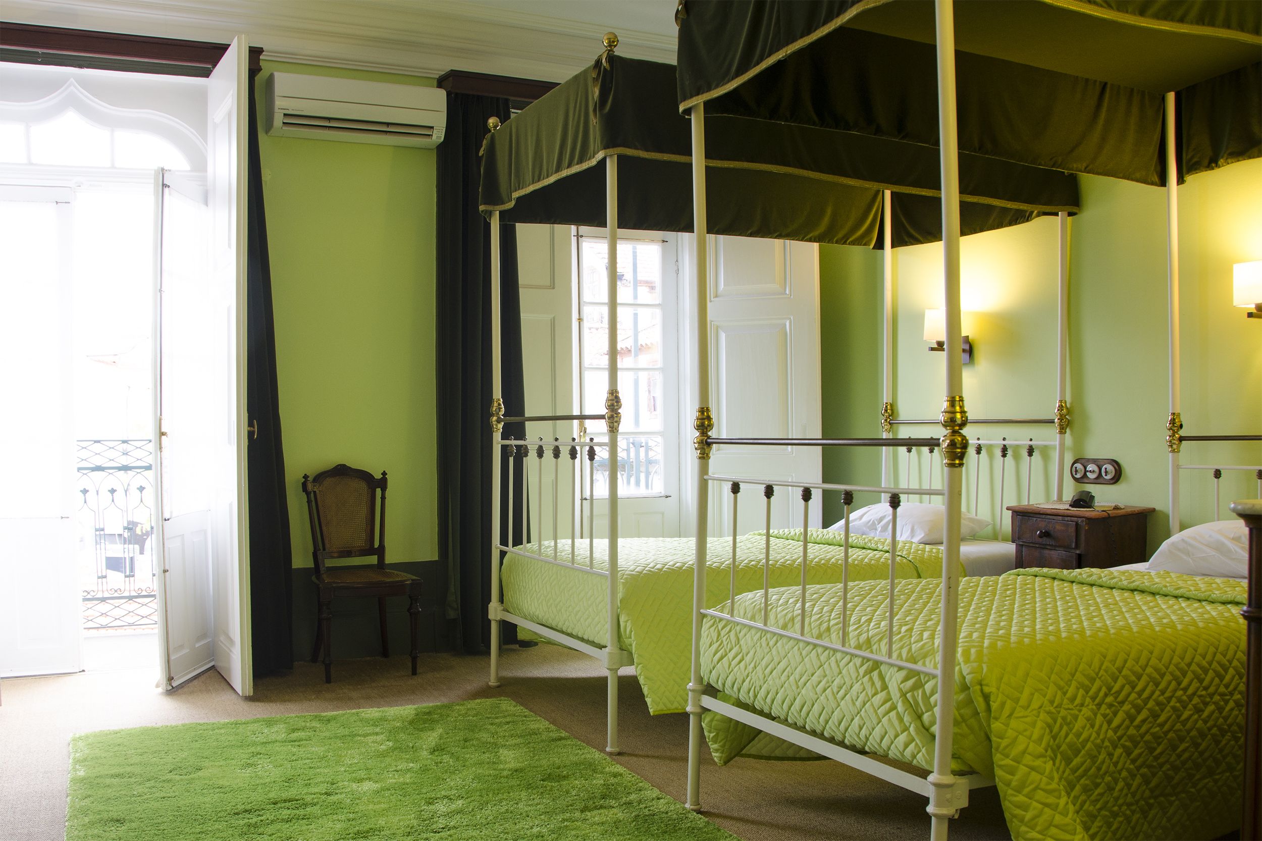 A four-poster bed with green bedding, a wooden chair, and a nightstand with a lamp.