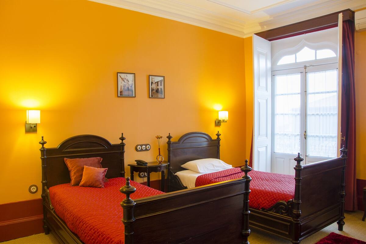 A double bed with a red bedspread, two wall-mounted lamps, and a window with curtains.