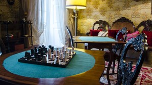 A room with a chessboard on a wooden table, a lamp, ornate chairs, and a patterned wallpaper background.