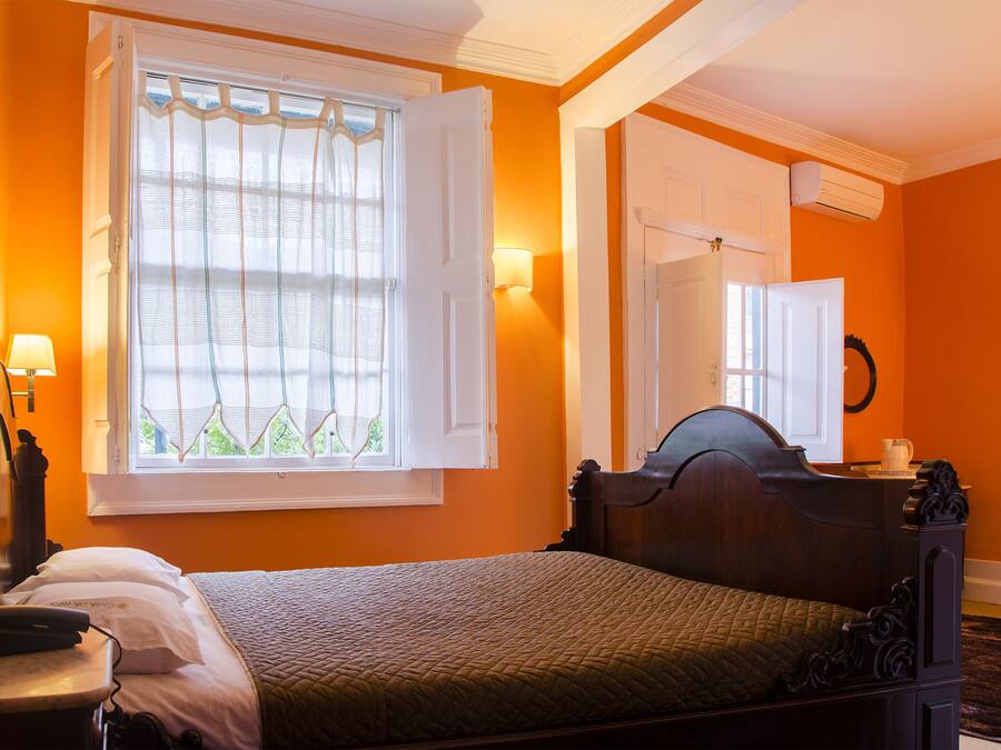 A bedroom with a bed, a window with curtains, a bedside lamp, a mirror, and an air conditioning unit.