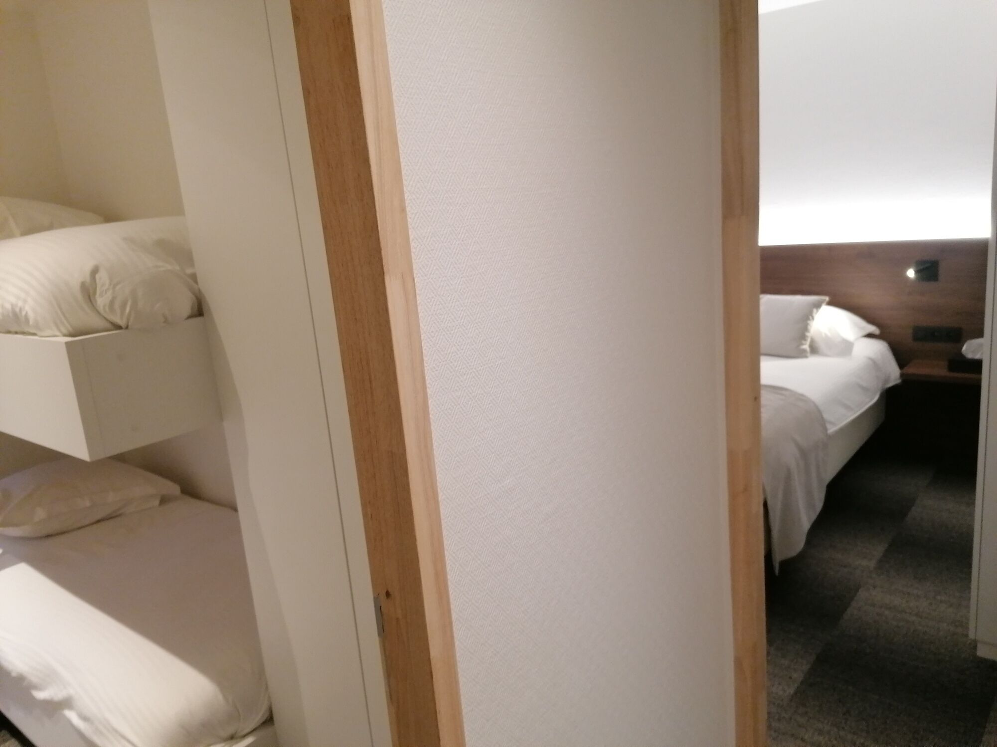 Quarto de hotel com beliche, uma cama de solteiro e mesa de cabeceira com abajur.