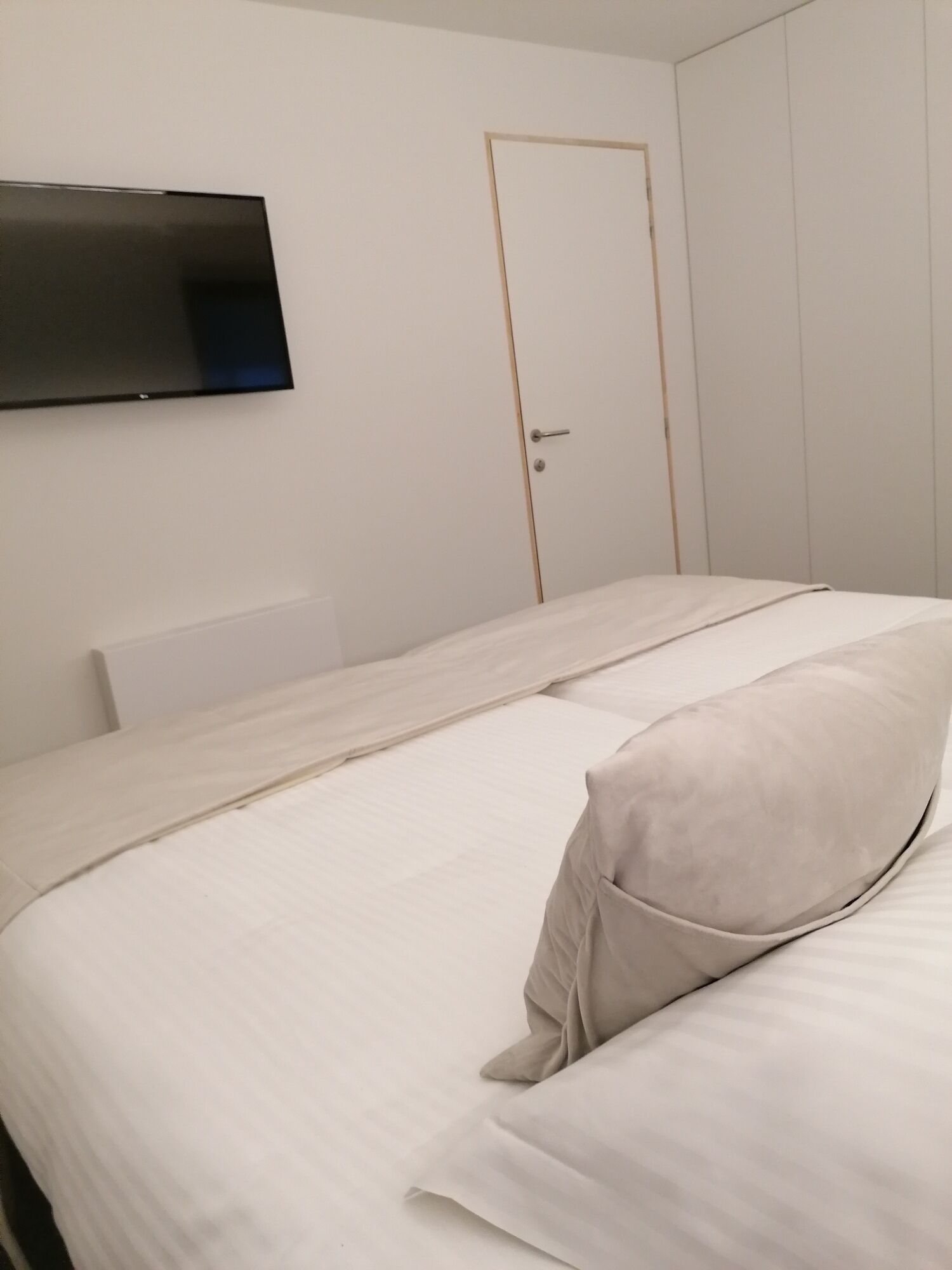 A minimalist hotel room with a flat-screen TV mounted on the wall, a single bed with a light-colored duvet and pillow, and a closed door.