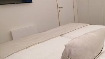 A minimalist hotel room with a flat-screen TV mounted on the wall, a single bed with a light-colored duvet and pillow, and a closed door.