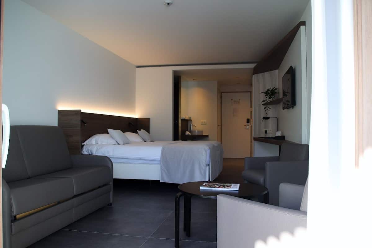 A modern hotel room with a bed, sofa, and armchair, a small table, and a TV.