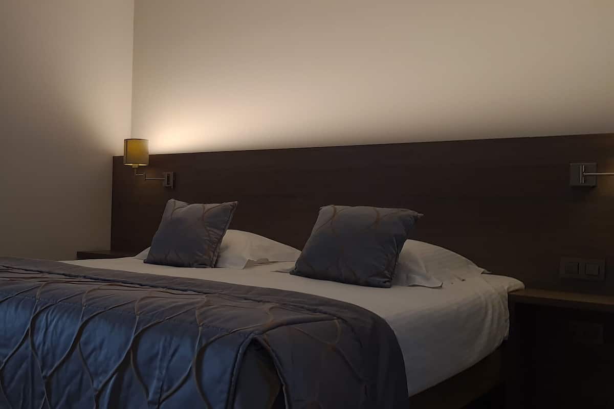 A hotel room with a bed, a nightstand, a wall-mounted TV, and a wall-mounted lamp.