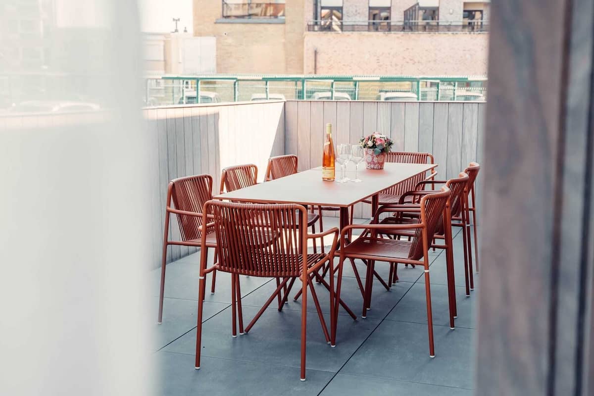 A rooftop terrace with a rectangular table and six wicker chairs, a bottle of wine, and a vase with flowers.