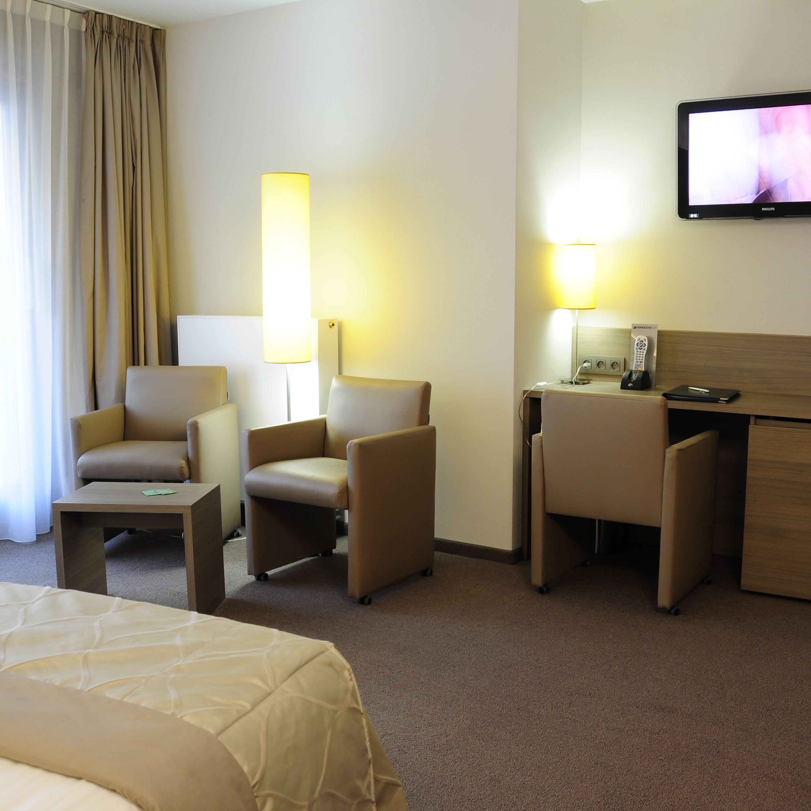 Ein Hotelzimmer mit einem Bett, zwei Sesseln, einem Schreibtisch, einem Fernseher und zwei Lampen.