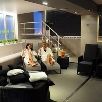 A modern lounge area with two people seated on cushioned chairs, a staircase, and a black armchair.