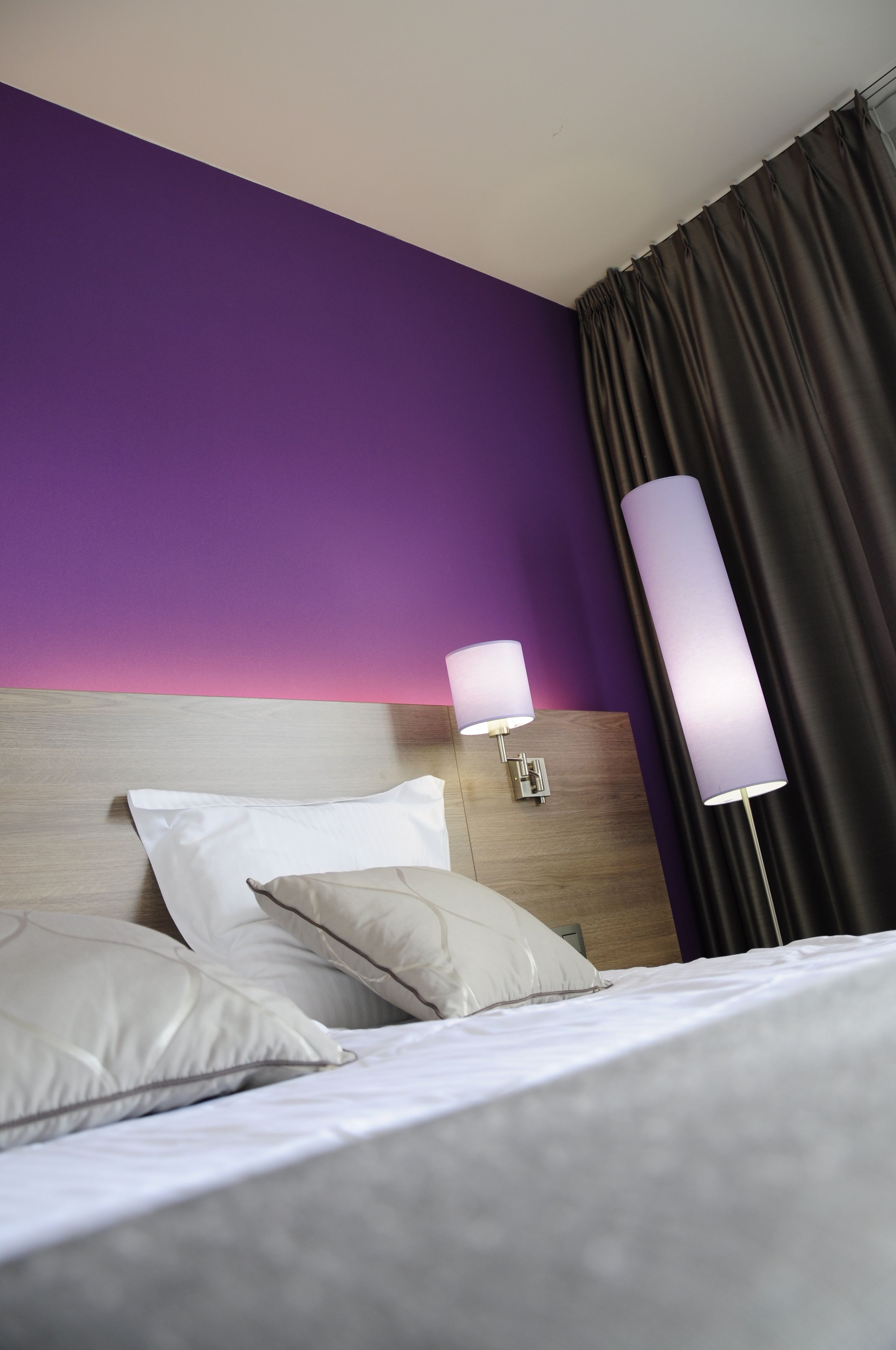 Een hotelkamer met een bed, kussens, een hoofdeinde, een wandlamp en een gordijn.