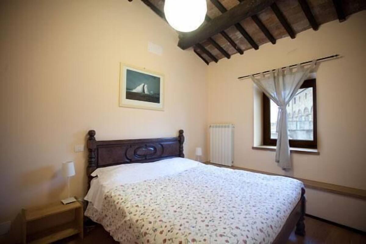 Une chambre à coucher avec un lit en bois, une table de chevet, une fenêtre avec des rideaux et un radiateur.