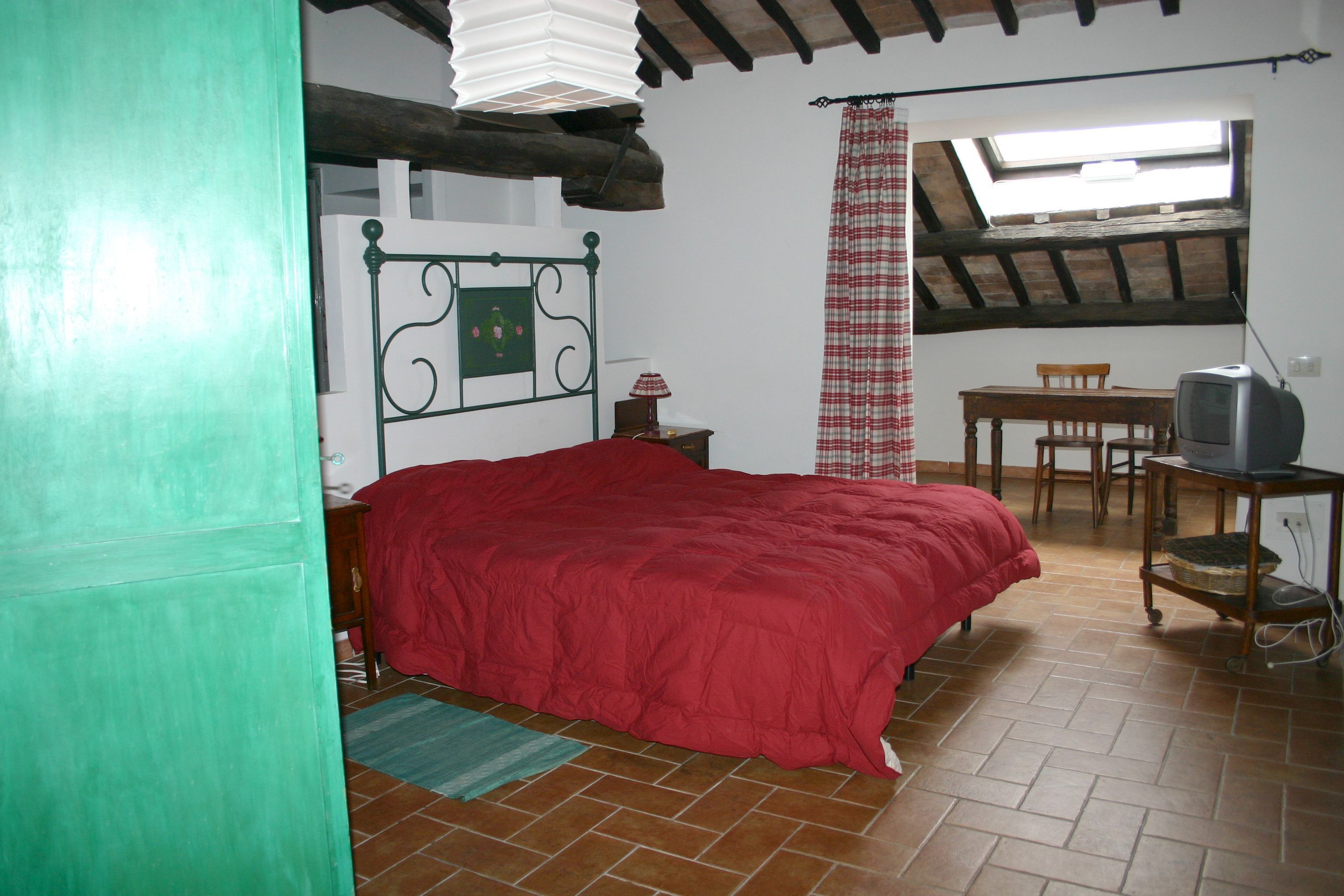 Un dormitorio con una cama roja, una mesa de madera con un televisor, una ventana con vistas, y una puerta verde.