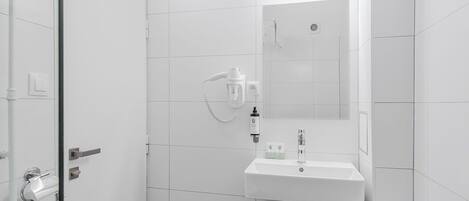 A modern bathroom with a white sink, mirror, and shower. There is a toilet paper holder and a small trash bin.