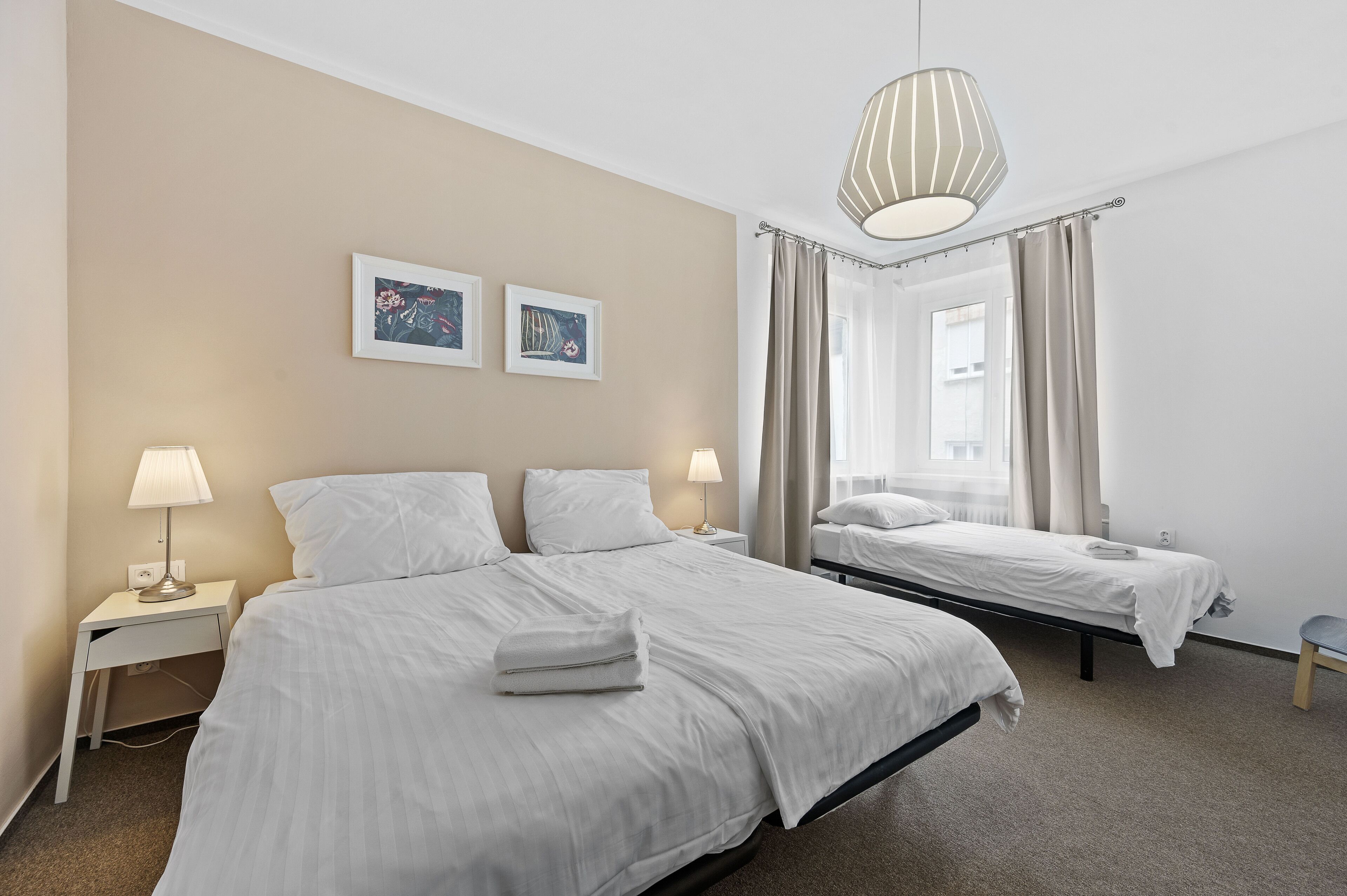 A hotel room with two beds, a nightstand with a lamp, a window with curtains, and two framed pictures on the wall.