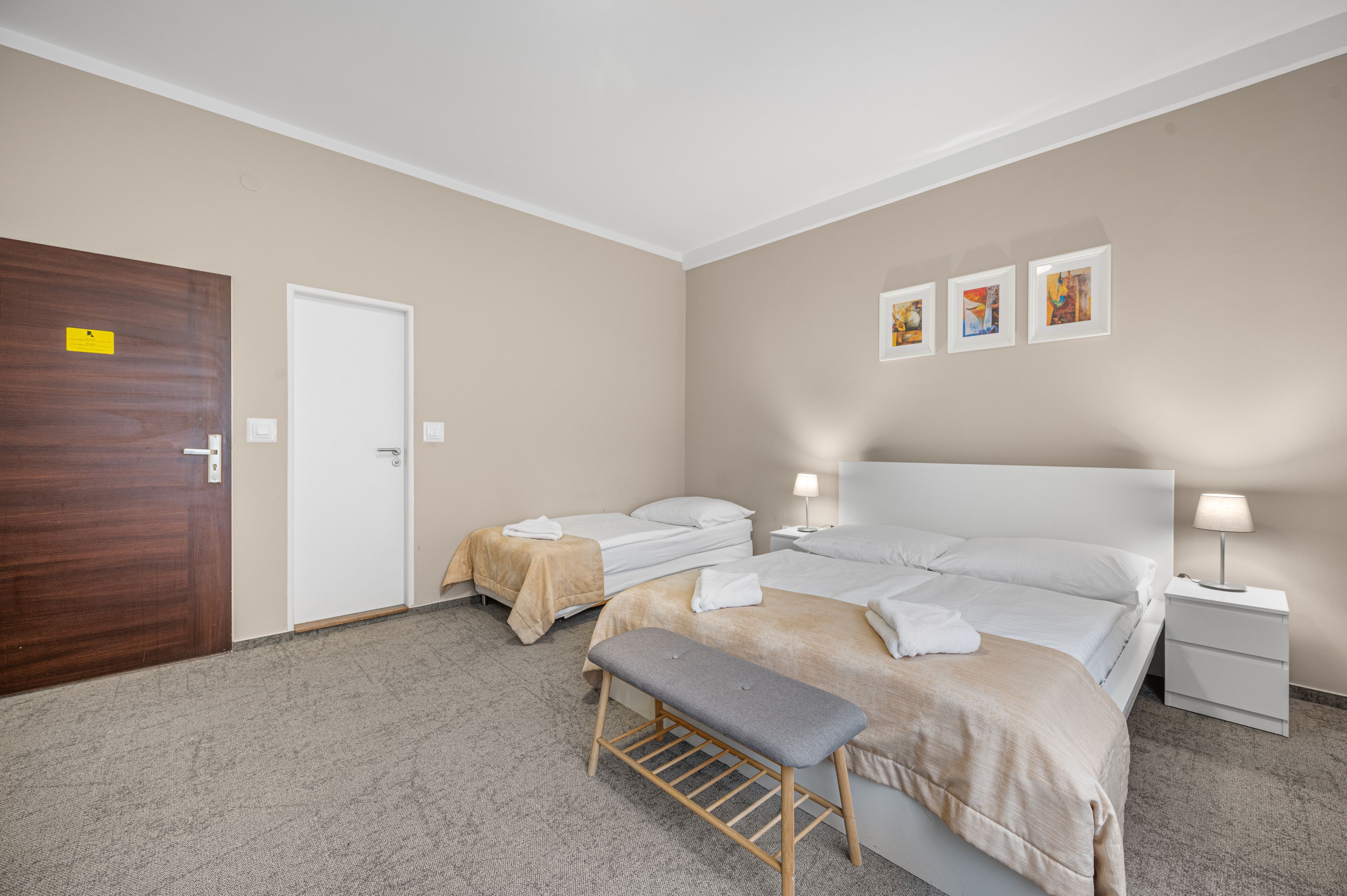 Ein Hotelzimmer mit zwei Einzelbetten, einer Bank, einem Nachttisch mit Lampe und einer Tür mit einem Schild.