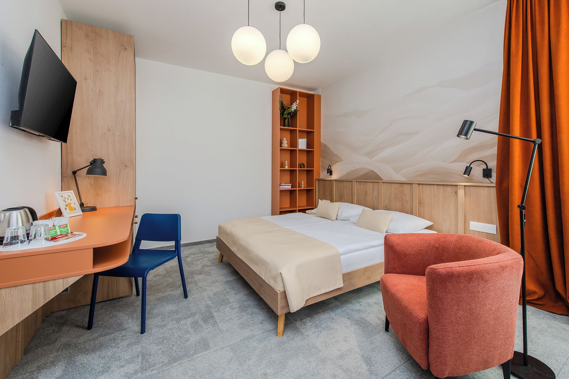 A hotel room with a bed, a desk with a chair, a TV, a bookshelf, and a lamp.