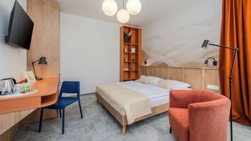 A hotel room with a bed, a desk with a chair, a TV, a bookshelf, and a lamp.